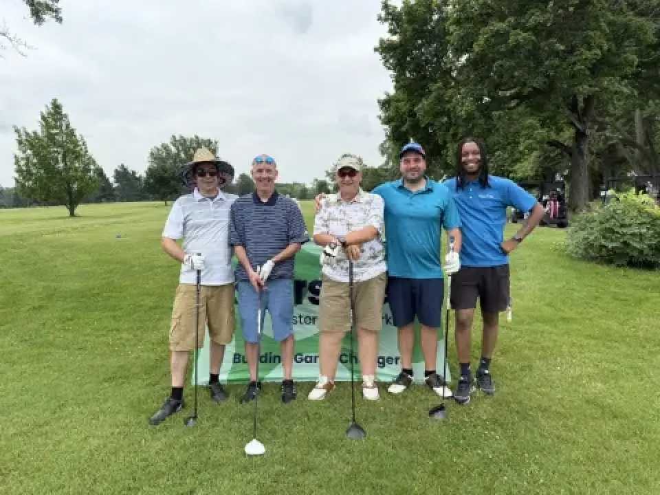 Five people with golf clubs pose together on a grassy golf course in front of a sign.