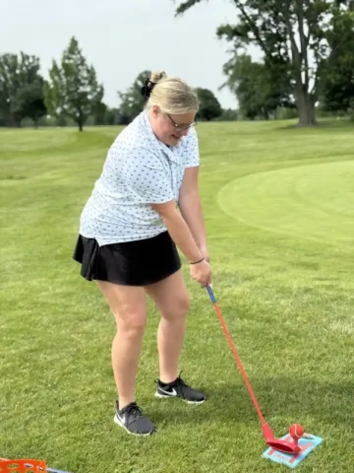 Person in glasses and golf attire prepares to putt a red ball on grass near a putting green.