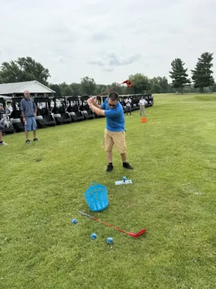 A man in blue swings a large red plastic golf club on a grassy field near golf carts.