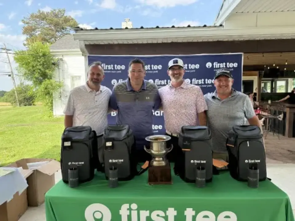 Four men stand behind golf prizes and a trophy, smiling in front of a first tee banner.