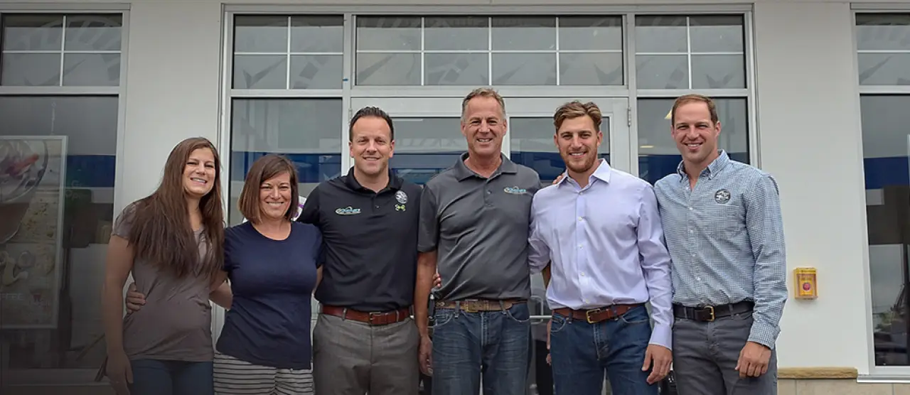 Six people standing together and smiling in front of a building with glass doors and windows.
