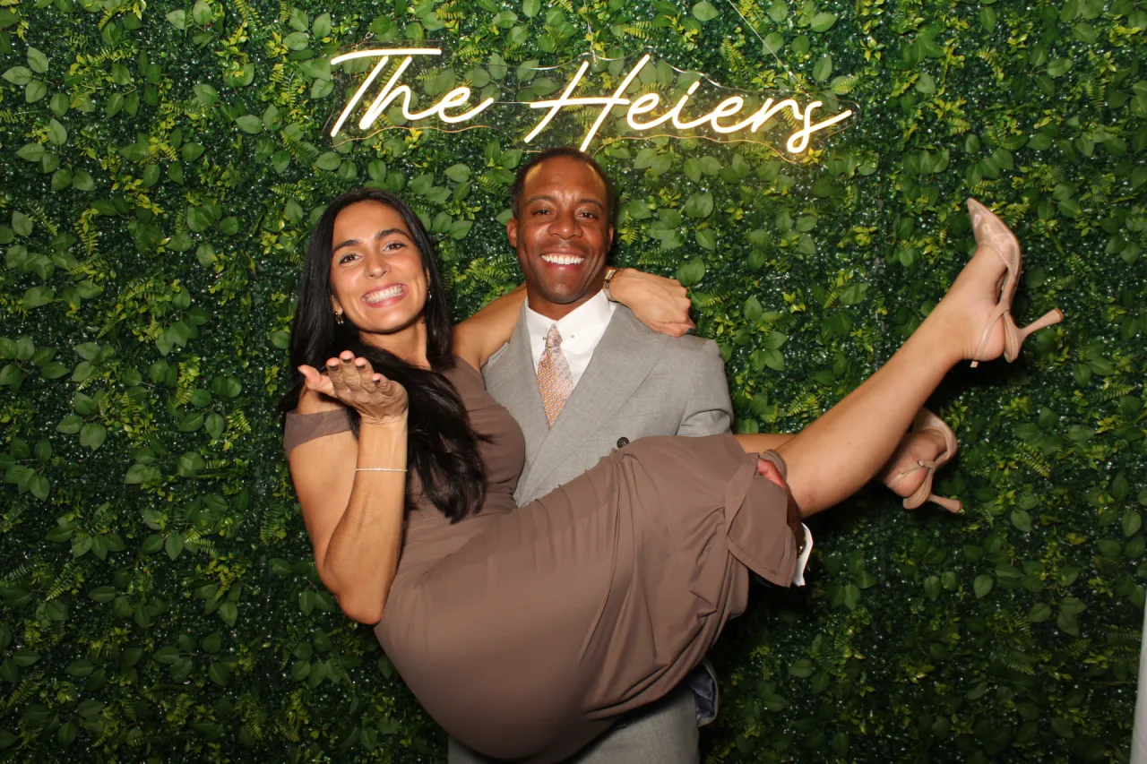 Couple posing in front of the Hedge Over Heels greenery wall with a custom neon sign