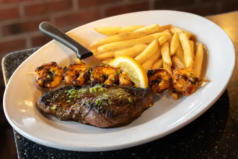 Grilled steak, shrimp skewer, fries, and a lemon wedge on a white plate with a knife.