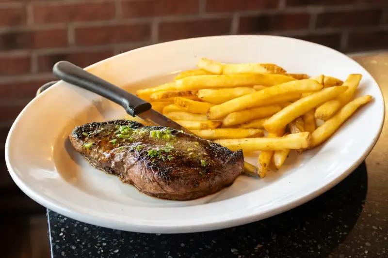 A plate with a grilled steak, French fries, and a knife on a white dish.