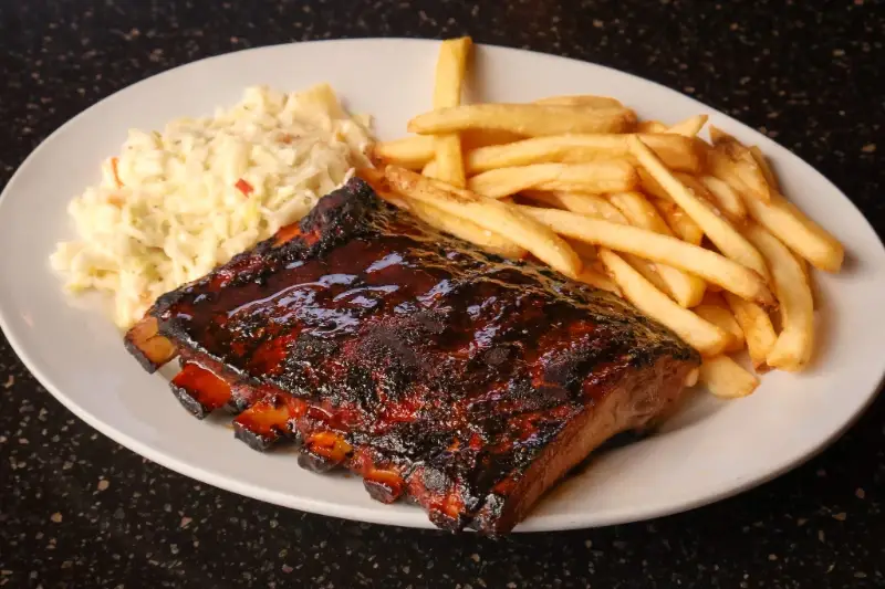 Barbecued ribs with French fries and coleslaw served on a white plate against a dark background.