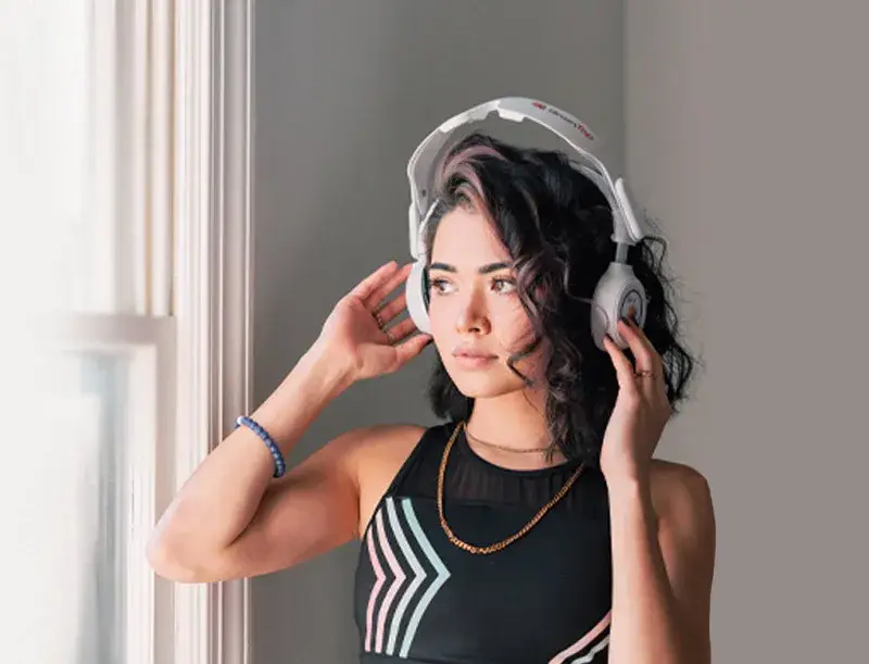 Woman with headphones stands near a window, looking outside while holding the headphones.