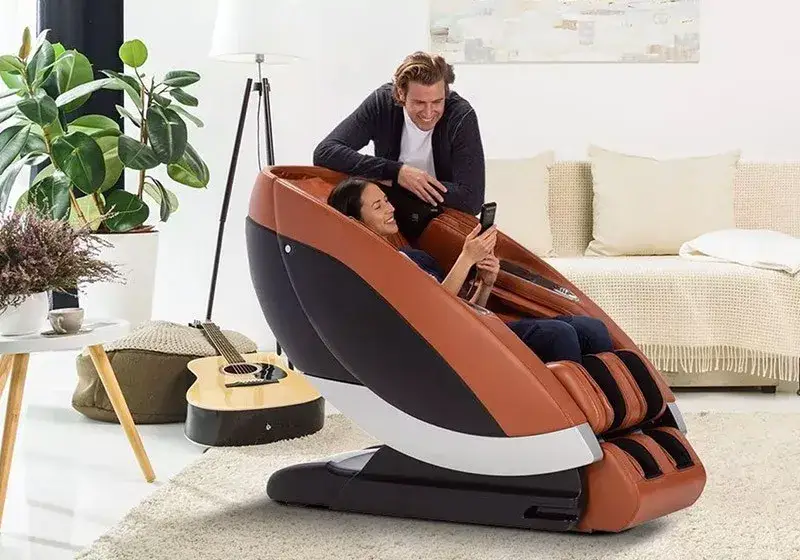 Woman relaxing in a massage chair while a man smiles and leans over her in a cozy living room.